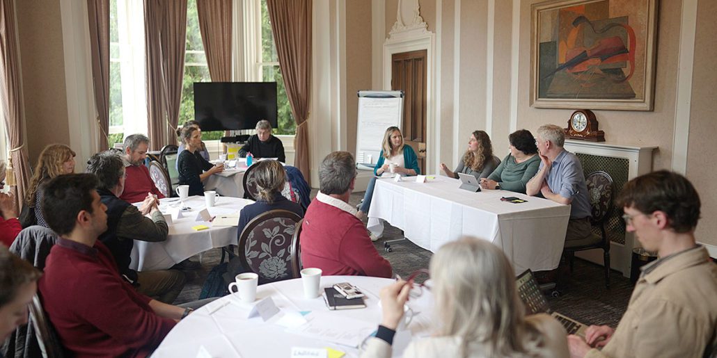 A group of people seated around round tables in a conference room with tall windows and elegant curtains. At the front, a panel of five individuals sits behind a rectangular table, engaging in discussion. The room features a large wall-mounted painting and an antique clock. Participants are actively listening and taking notes, creating an atmosphere of focused discussion and collaboration.